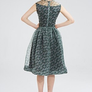May include: A sleeveless, knee-length dress with a floral pattern. The dress features a sheer overlay and a fitted bodice that flares into a full skirt. The color is a muted green with darker green floral accents. The model is wearing black high heels.