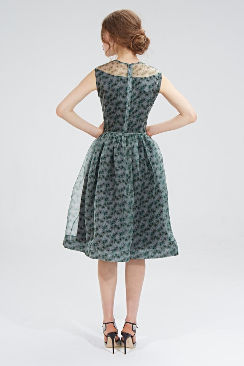 May include: A sleeveless, knee-length dress with a floral pattern. The dress features a sheer overlay and a fitted bodice that flares into a full skirt. The color is a muted green with darker green floral accents. The model is wearing black high heels.
