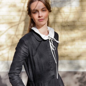 May include: A woman wearing a black linen dress with a white peter pan collar and a white ribbon tied at the neck. The dress has a fitted bodice and a full skirt. The woman is standing in front of a brick wall.