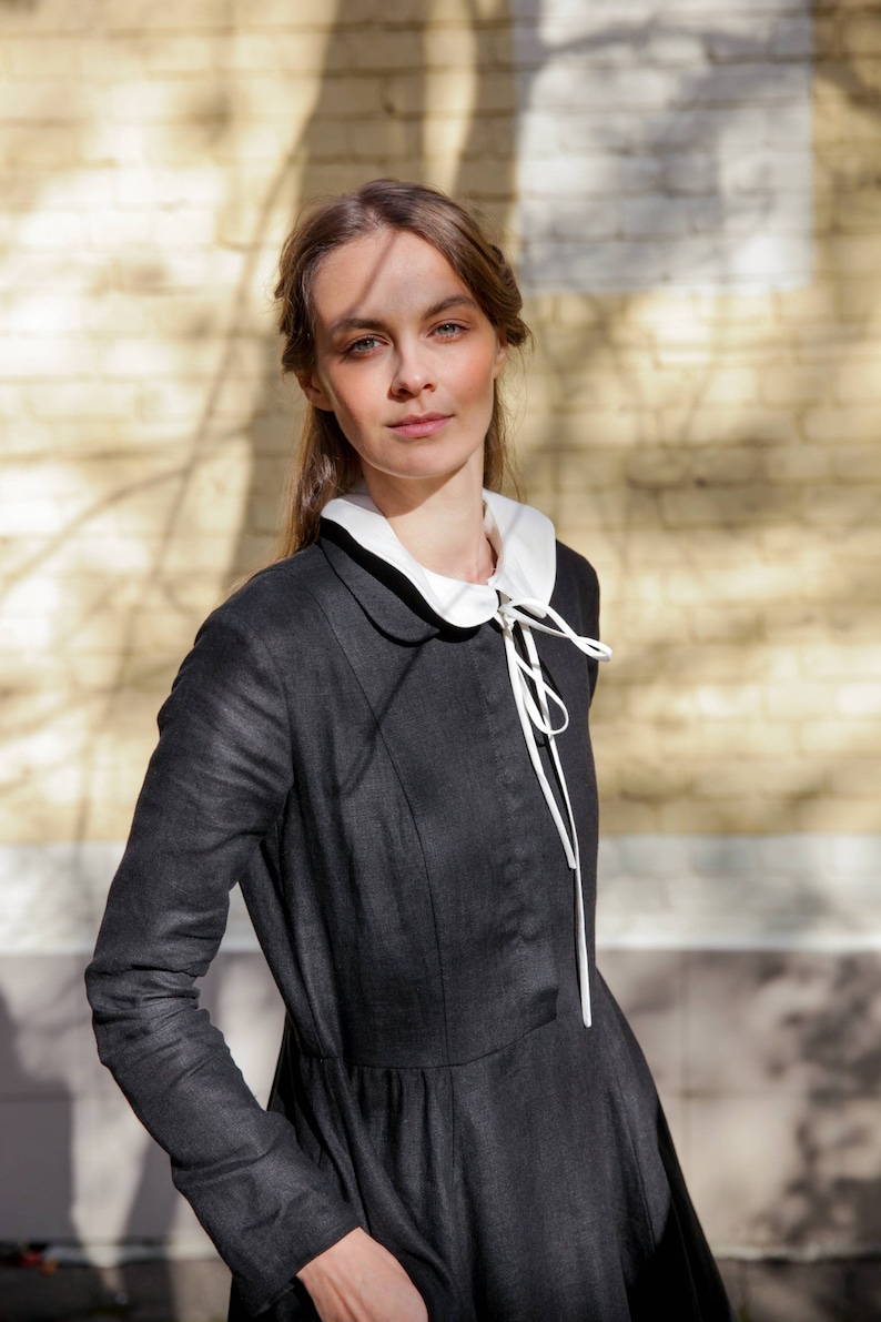 May include: A woman wearing a black linen dress with a white peter pan collar and a white ribbon tied at the neck. The dress has a fitted bodice and a full skirt. The woman is standing in front of a brick wall.
