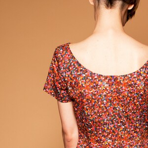 May include: A woman wearing a red, short-sleeved dress with a colorful confetti print. The dress has a scoop neckline and a back zipper.