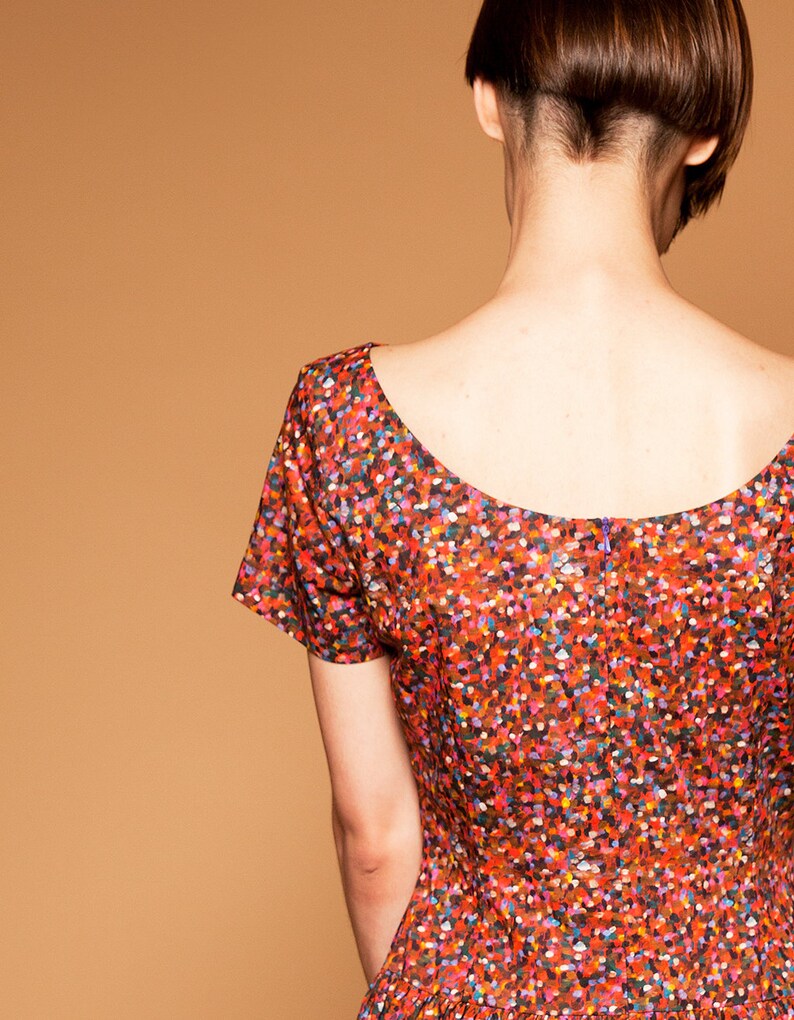 May include: A woman wearing a red, short-sleeved dress with a colorful confetti print. The dress has a scoop neckline and a back zipper.