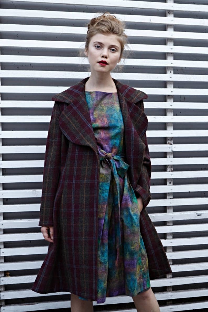 May include: A woman wearing a long, plaid coat over a colorful, tie-dye dress. The coat has a large collar and a tie at the waist. The woman is standing in front of a white slatted wall.