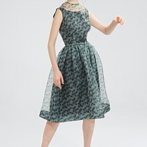 May include: A sleeveless, knee-length dress with a floral pattern in shades of green and gray. The dress features a sheer overlay on the skirt and neckline. The model is wearing black high-heeled sandals.