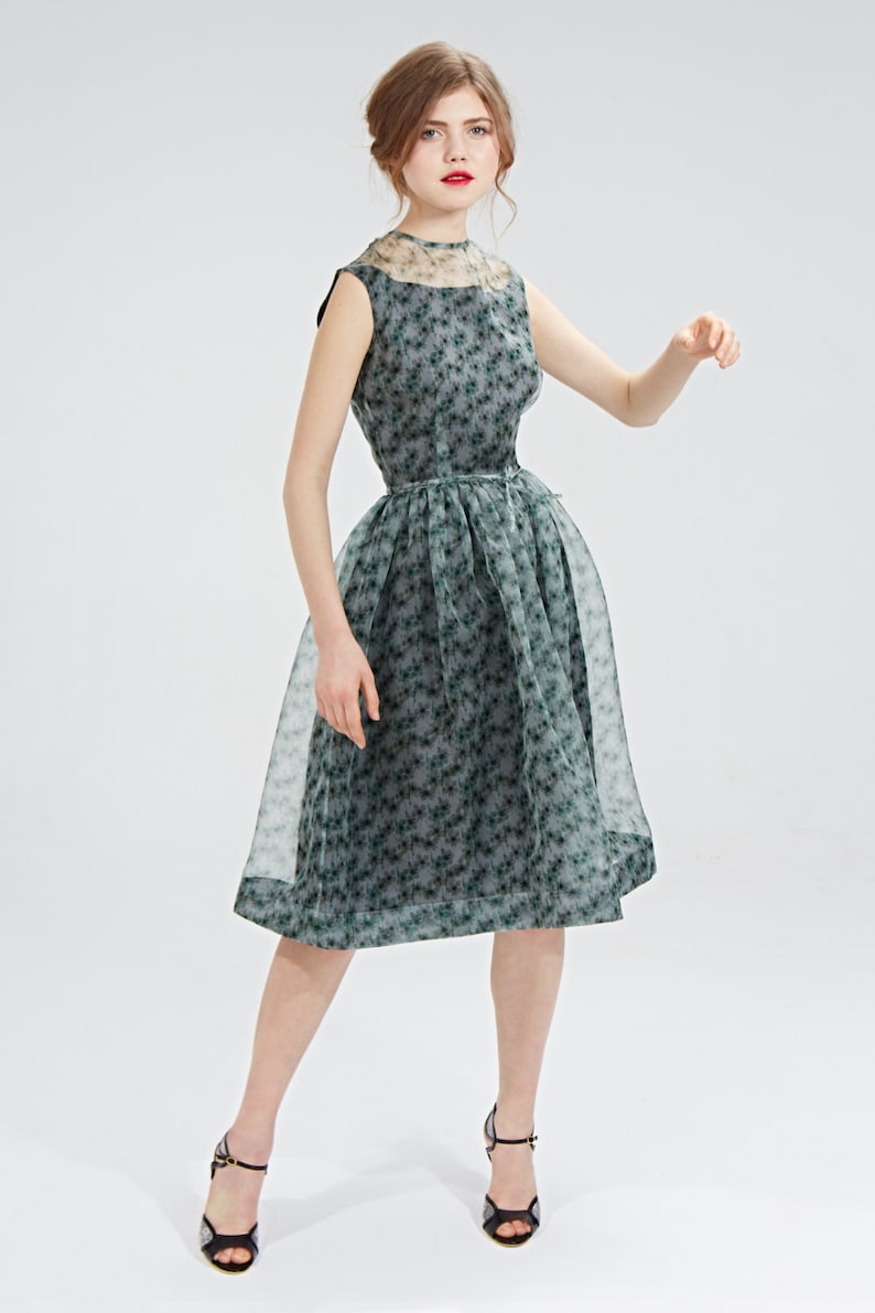 May include: A sleeveless, knee-length dress with a floral pattern in shades of green and gray. The dress features a sheer overlay on the skirt and neckline. The model is wearing black high-heeled sandals.