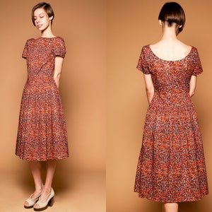 May include: A woman wearing a short-sleeved, knee-length dress with a red, orange, and yellow floral print. The dress has a scoop neckline and a fitted bodice. The woman is wearing her hair in a short, choppy style.