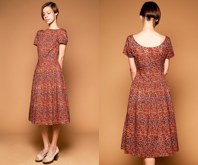 May include: A woman wearing a short-sleeved, knee-length dress with a red, orange, and yellow floral print. The dress has a scoop neckline and a fitted bodice. The woman is wearing her hair in a short, choppy style.