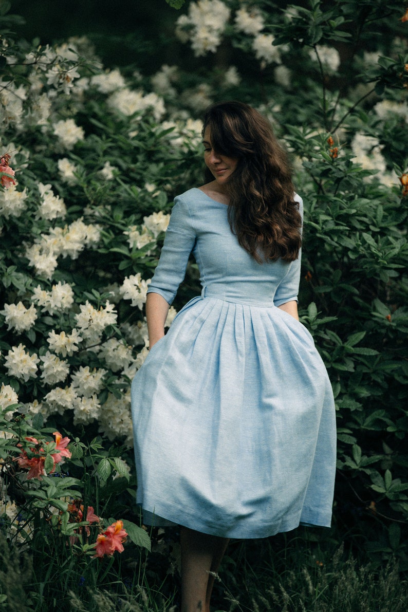 A woman wearing a light blue linen midi dress poses elegantly amidst a colorful array of blooming flowers. She iskeeping herhands in thepockets.