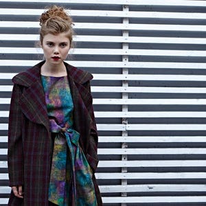 May include: A woman wearing a long-sleeved, plaid coat with a hood over a colorful, patterned dress. She is standing in front of a white slatted wall.