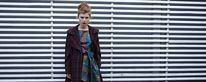May include: A woman wearing a long-sleeved, plaid coat with a hood over a colorful, patterned dress. She is standing in front of a white slatted wall.