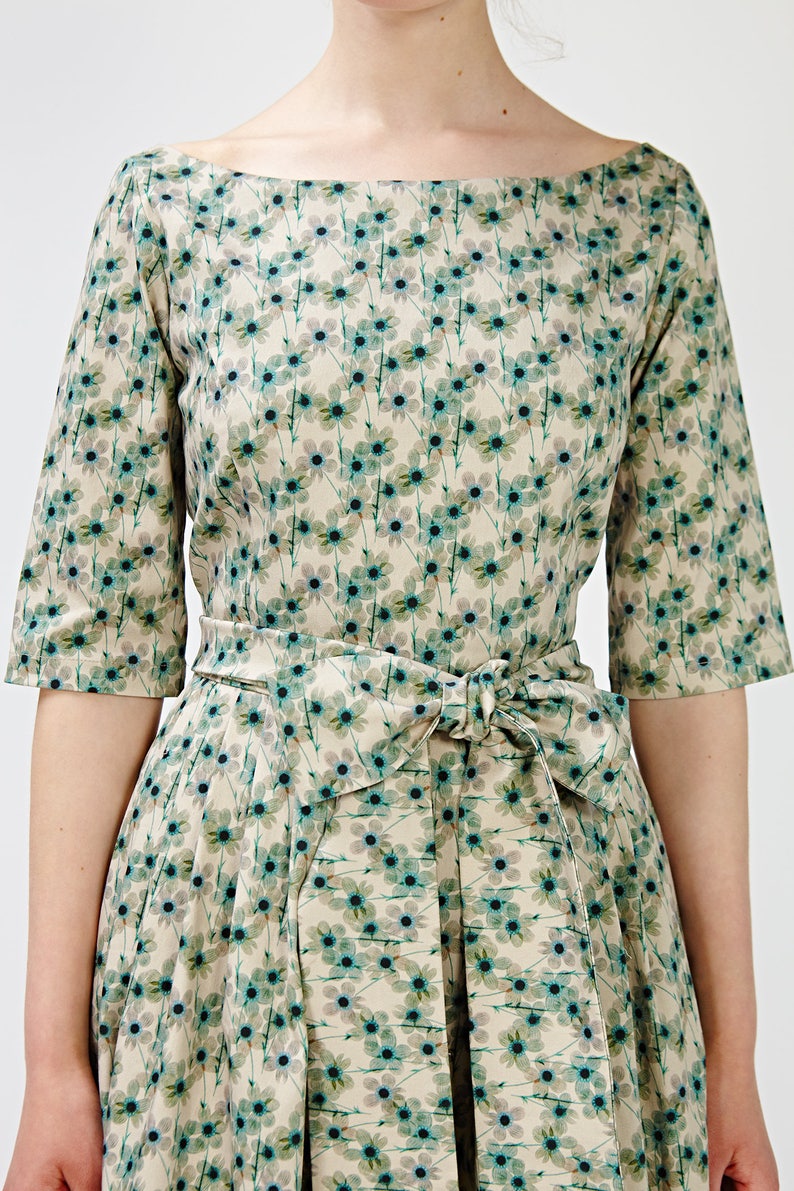 May include: A beige dress with a floral print featuring small blue flowers. The dress has a boat neck and a self-tie sash.