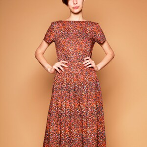 May include: A woman wearing a short-sleeved, knee-length dress with a red, orange, and green floral print. The dress has a boat neck and a fitted bodice.