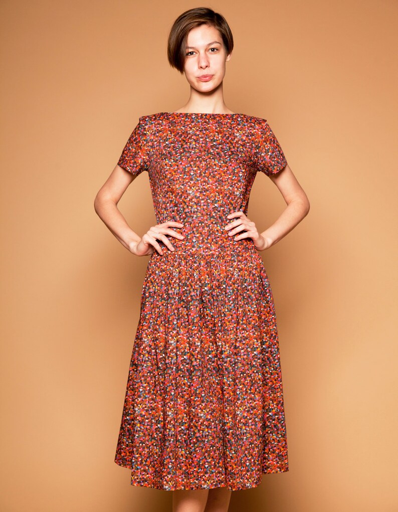 May include: A woman wearing a short-sleeved, knee-length dress with a red, orange, and green floral print. The dress has a boat neck and a fitted bodice.