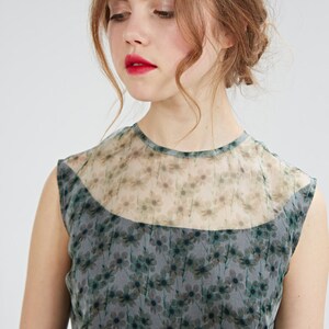 May include: A sleeveless dress featuring a floral pattern in shades of gray and green. The dress has a sheer neckline and a fitted bodice. The model is wearing the dress, with red lipstick and her hair styled up.