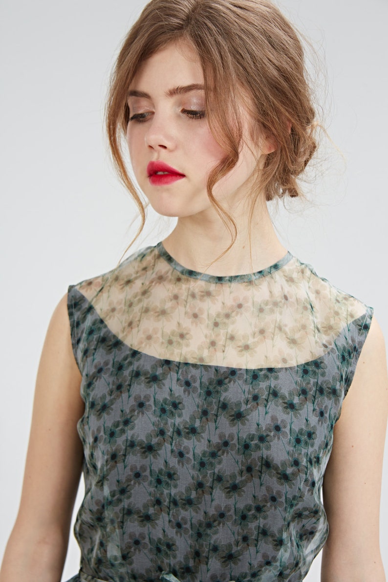 May include: A sleeveless dress featuring a floral pattern in shades of gray and green. The dress has a sheer neckline and a fitted bodice. The model is wearing the dress, with red lipstick and her hair styled up.