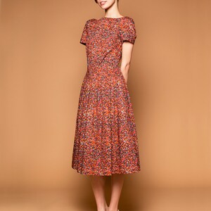May include: A woman wearing a short-sleeved, red, orange, and blue floral print dress with a white background. She is wearing white heeled shoes.