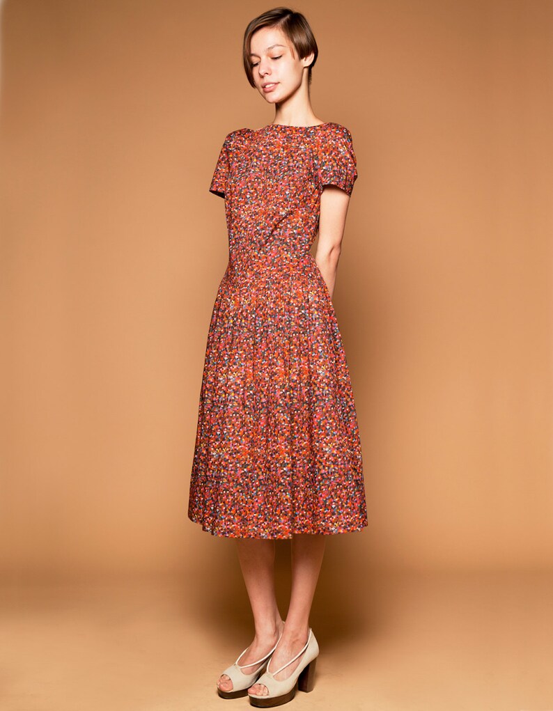 May include: A woman wearing a short-sleeved, red, orange, and blue floral print dress with a white background. She is wearing white heeled shoes.