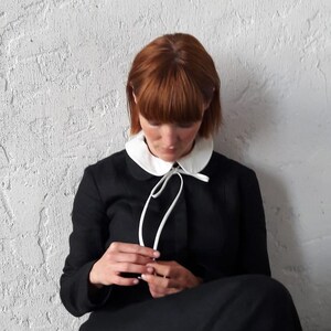 May include: A woman wearing a black dress with a white Peter Pan collar and a white ribbon tied at the neck. The collar has a small bow at the center.