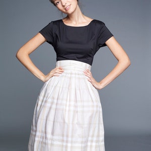 May include: A woman wearing a black short-sleeved top and a white and beige plaid skirt.