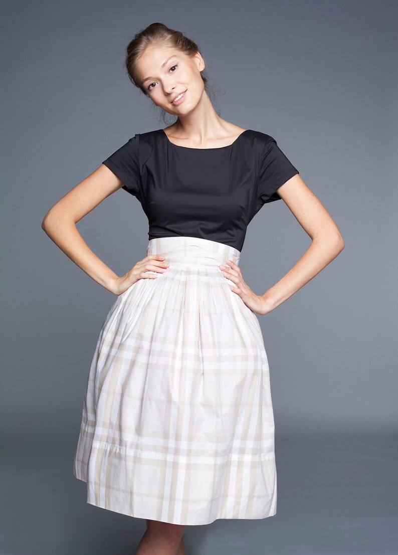 May include: A woman wearing a black short-sleeved top and a white and beige plaid skirt.