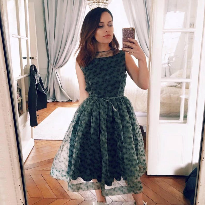 May include: A sleeveless, knee-length dress in a dark green hue, adorned with a delicate floral pattern. The dress features a sheer overlay, a fitted bodice, and a flared skirt. A thin belt cinches the waist. The dress is worn with sneakers.