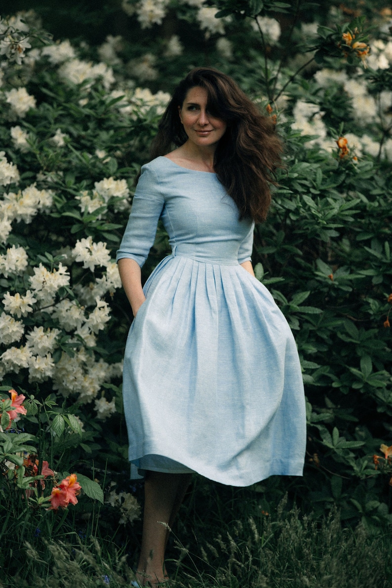 A woman in a light blue linen midi dress stands gracefully in front of a vibrant display of flowers.
