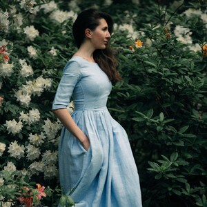 A woman in a blue midi dress wiht tucked skirt stands in front of white azalea bush. She has her hands in her pockets.