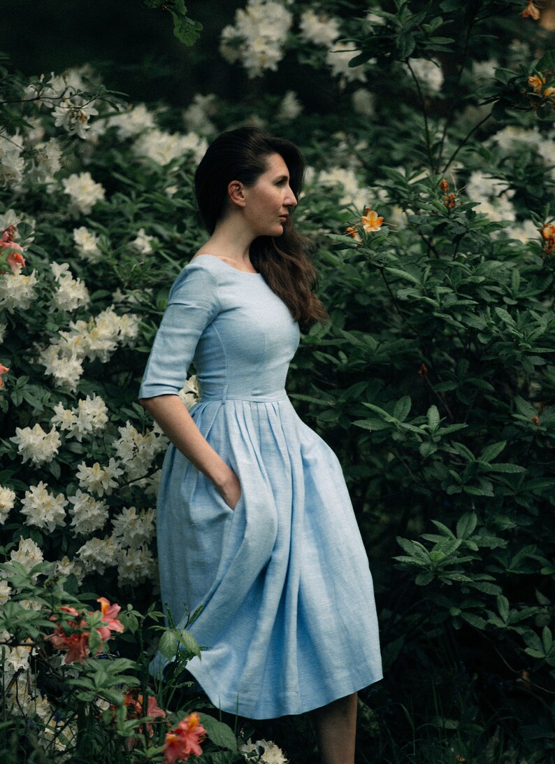 A woman in a blue midi dress wiht tucked skirt stands in front of white azalea bush. She has her hands in her pockets.