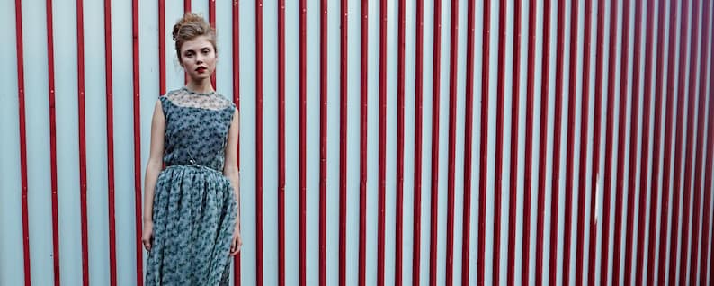 May include: A woman wearing a gray floral print dress with a sheer neckline and a matching skirt. The dress is cinched at the waist with a silver belt. The background features a red and white striped wall.