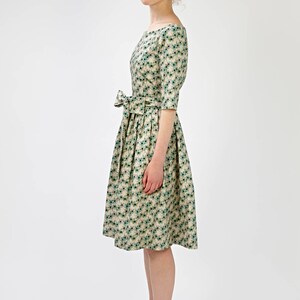 May include: A beige dress with a green floral pattern and a bow at the waist. The dress has a boat neck and three-quarter sleeves. The model is wearing brown heeled sandals.