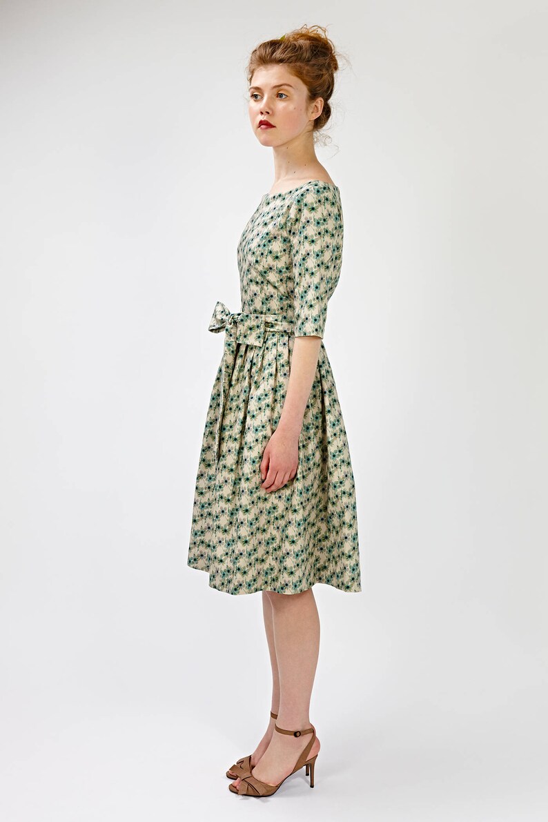 May include: A beige dress with a green floral pattern and a bow at the waist. The dress has a boat neck and three-quarter sleeves. The model is wearing brown heeled sandals.