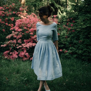 A woman in a blue swing midi dress with three-quarter sleeves stands in front of pink azalea bush, wearing stylish heels.