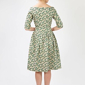 May include: A beige dress with a green floral pattern and a boat neckline. The dress has a fitted bodice and a full skirt. The model is wearing brown heeled sandals.