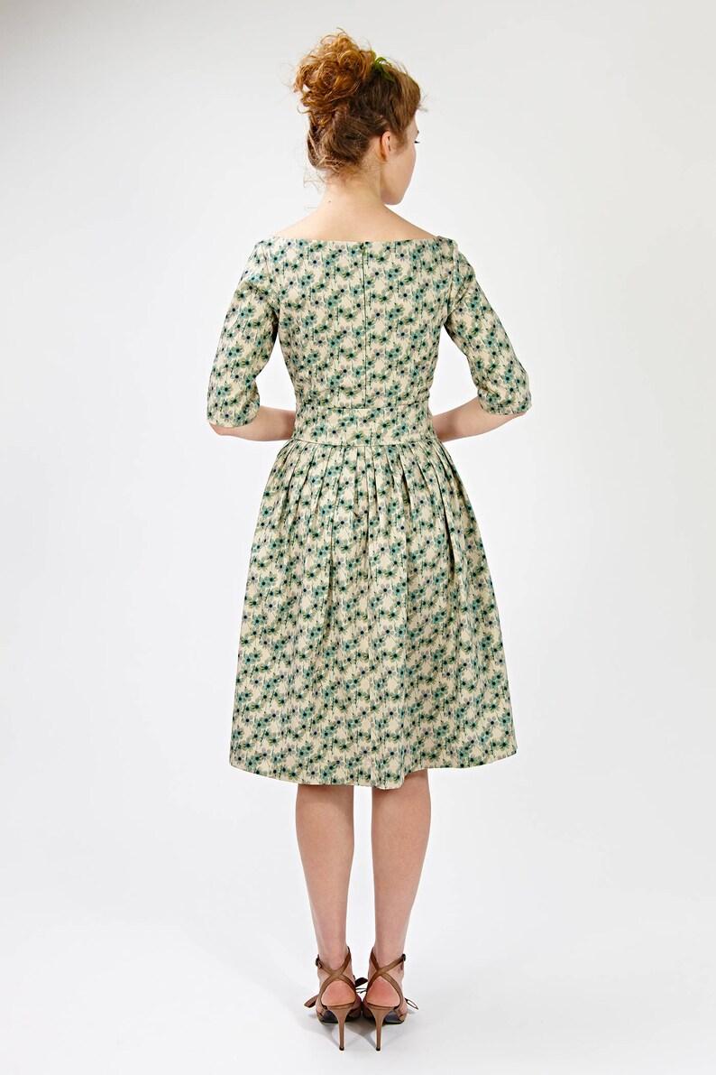 May include: A beige dress with a green floral pattern and a boat neckline. The dress has a fitted bodice and a full skirt. The model is wearing brown heeled sandals.