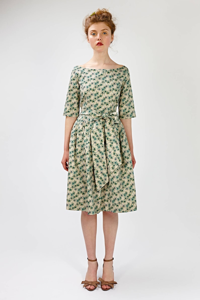 May include: A woman wearing a beige and green floral print dress with a matching sash tied at the waist. She is wearing brown heeled sandals with a bow detail.