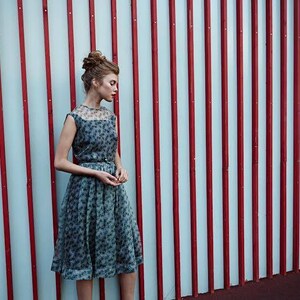 May include: A woman wearing a vintage-style dress with a floral pattern. The dress is a soft blue-gray color and has a sheer overlay on the bodice. The background features a red and white striped wall.