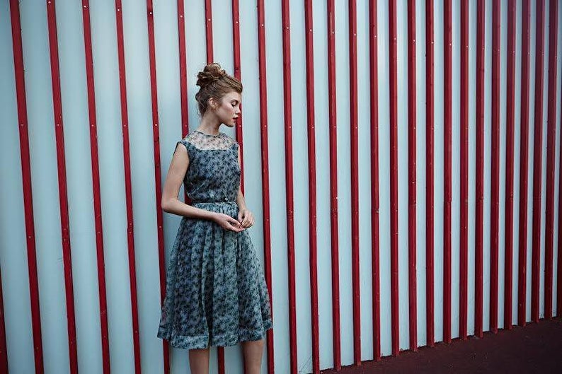 May include: A woman wearing a vintage-style dress with a floral pattern. The dress is a soft blue-gray color and has a sheer overlay on the bodice. The background features a red and white striped wall.
