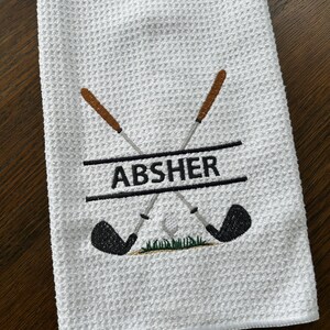 Op de afbeelding: Witte golfhanddoek met een wafelweefseltextuur. Geborduurd met gekruiste golfclubs, groen gras en de naam "ABSHER" in zwart. De golfclubs hebben bruine handvatten en zwarte clubhoofden.
