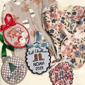 Op de afbeelding: Geborduurde kerstornamenten met namen en data, naast baby pyjama's. Een ornament heeft een kerstman-ontwerp, terwijl andere namen weergeven zoals "Max", "Bennett" en "Noah". De pyjama's hebben koekjes- en bloempatronen en bloemen.