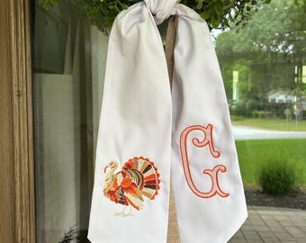 May include: White fabric door hanger with a colorful embroidered turkey and an orange monogram letter "G". The door hanger is tied to a green wreath, creating a festive fall decoration.