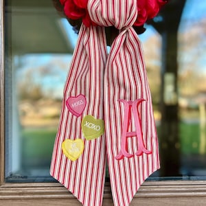 Op de afbeelding: Een rood-witte gestreepte stoffen strik met een grote roze letter "A" en drie hartvormige patches. De patches hebben de woorden "HUGS", "XOXO" en "BE MINE" geborduurd. De strik is bevestigd aan een rode bloemenkrans.