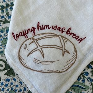 May include: White linen kitchen towel with an embroidered bread loaf design and the phrase "loafing him was bread" in burgundy thread. The towel is folded, showing a corner with the design.