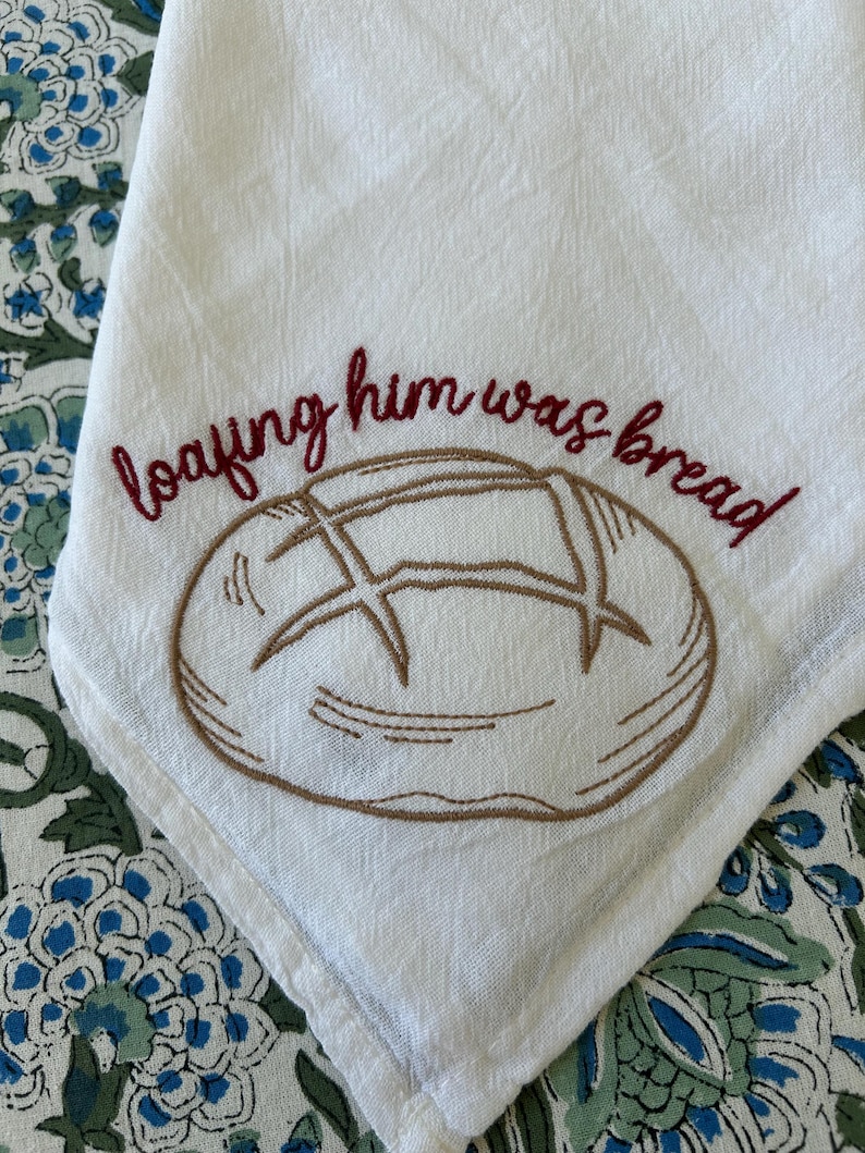 Op de afbeelding: Witte linnen keukendoek met een geborduurd broodontwerp en de zin "loafing him was bread" in bordeauxrode draad. De handdoek is gevouwen en toont een hoek met het ontwerp.