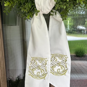 Op de afbeelding: Witte decoratieve sjerp met een geborduurd goud en zwart letter "S" ontwerp. De sjerp hangt aan een groene krans. De sjerp is gemaakt van een zachte, vloeiende stof.