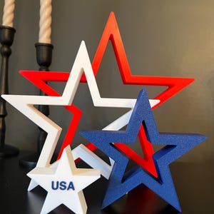 May include: A collection of star-shaped decorations in red, white, and blue. One small white star has the word "USA" printed on it. The other stars are larger and layered, with a mix of solid and open designs. These patriotic decorations are perfect for Independence Day.