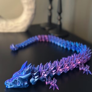 May include: A 3D-printed articulated dragon toy in shades of blue and purple. The dragon has a detailed, spiky design and is lying on a dark surface. The dragon's body is segmented, allowing for flexible posing.