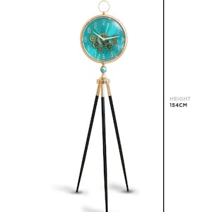 May include: A teal and gold floor clock with a steampunk design. The clock face is 35cm in diameter and features gears. The clock stands 154cm tall and has a 45cm base. The clock has a gold frame and black legs.