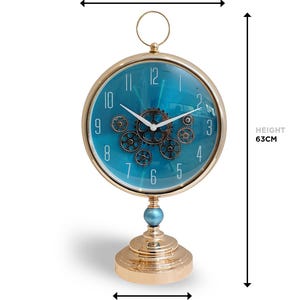 May include: A gold-colored, steampunk-style desk clock with a teal face and visible gears. The clock dial is 13.8 inches in diameter and the clock is 24.8 inches tall. The base is 7.7 inches wide. The clock has a round top and a decorative base.