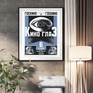 May include: A framed vintage movie poster with a black border. The poster features a large eye, the text "Kino Glaz 3", and other Cyrillic text. The color scheme is primarily blue, white, and black. The poster is hanging on a grey wall.