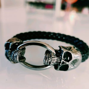 May include: A black leather bracelet with two silver skull charms. The bracelet has a silver ring clasp.